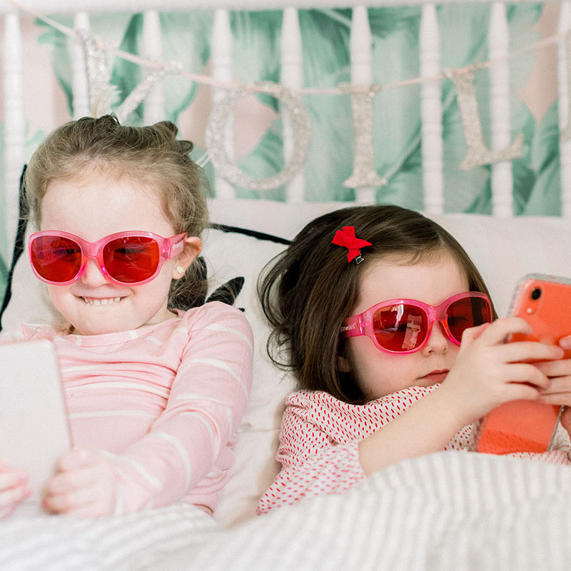 Two Girls playing on mobile devices while wearing TrueDark Kids Pink Frame Twilight Glasses
